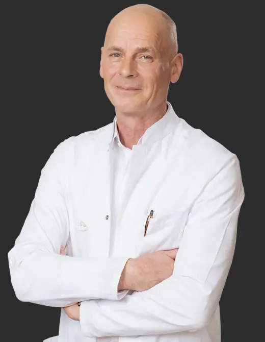 Ärztlich geführte Rehabilitation Porträt eines Arztes im weißen Kittel mit verschränkten Armen vor dunklem Hintergrund.